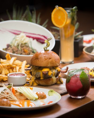 A wooden table in a bar with burger, fries, ketchup and cocktails on top